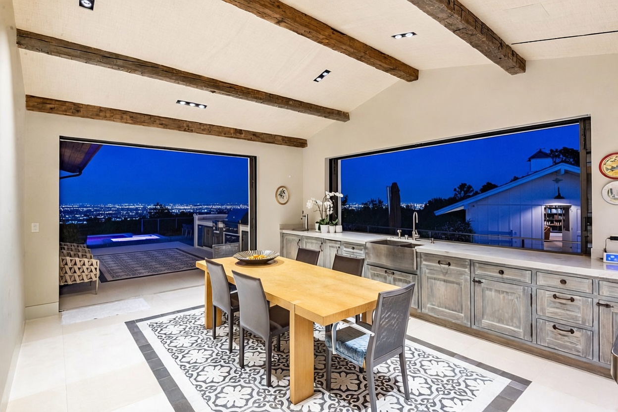 This open-concept kitchen and dining area features rustic wood ceiling beams and a large dining table set on a patterned tile rug. The space is defined by its expansive, wall-to-wall glass openings that provide a seamless transition to an outdoor patio and a panoramic view of the city lights at night. The kitchen is equipped with light-toned cabinetry, a farmhouse-style sink, and a clean, neutral aesthetic that emphasizes the indoor-outdoor connection.