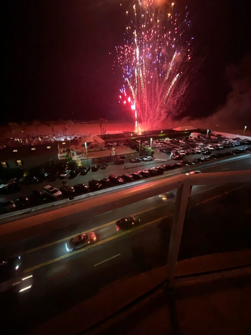 This nighttime view is captured from an elevated balcony, looking down over a busy street and a parking lot toward a beach. The scene is dramatically illuminated by a vibrant fireworks display exploding in the dark sky above the coastline. The perspective provides a clear vantage point of the surrounding neighborhood activity and the coastal atmosphere.