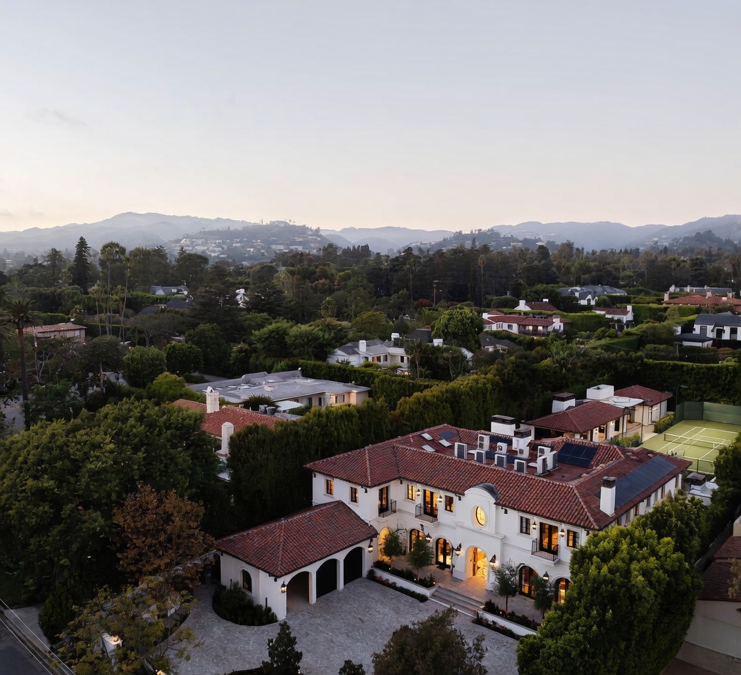 This high-angle aerial view captures a sprawling Mediterranean-style estate nestled within a lush, tree-lined neighborhood. The property features a multi-gabled terracotta tile roof, a gravel motor court with a detached garage, and a private tennis court visible on the right. The surrounding landscape is dense with mature greenery, set against a backdrop of rolling hills under a soft twilight sky.