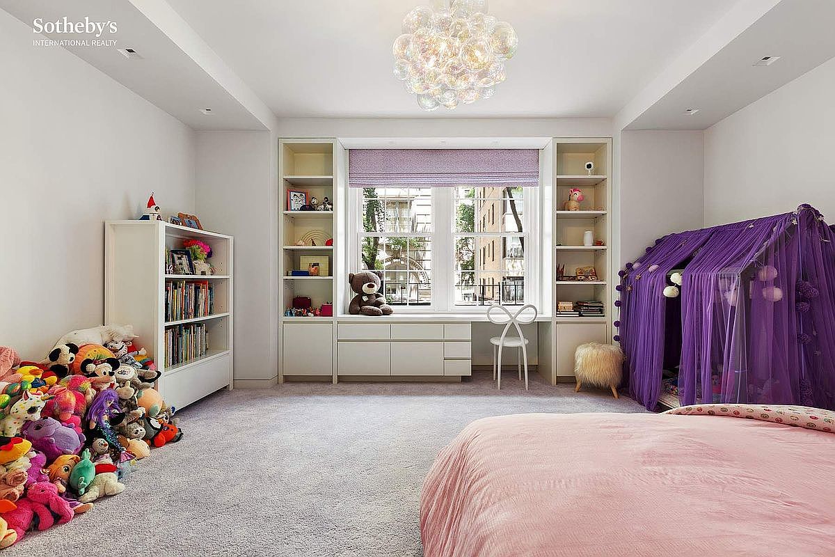 This bright and playful child's bedroom features a soft pink bed, a purple canopy play tent, and a large collection of stuffed animals. A built-in desk area is positioned beneath a window with lavender Roman shades, flanked by custom shelving units. The room is illuminated by a unique, multi-spherical glass chandelier and features neutral carpeting throughout.