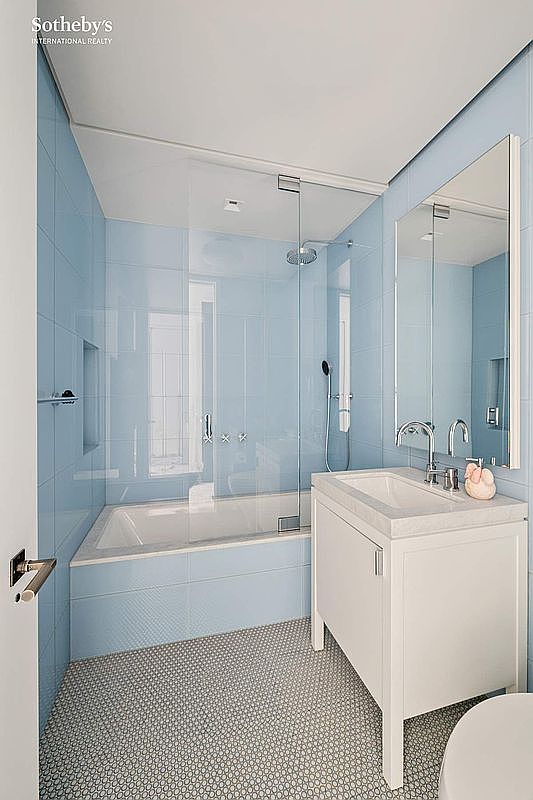 This bright, modern bathroom features light blue wall tiles and a distinctive penny-round mosaic floor. The space includes a glass-enclosed shower-tub combination and a white vanity with a chrome faucet and a tall, rectangular mirror. The perspective is a straight-on view from the doorway, highlighting the clean, minimalist aesthetic of the room.