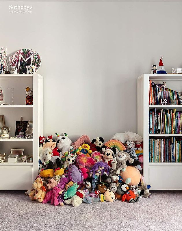 This image captures a child's bedroom space featuring two white, open-shelving bookcases flanking a central wall. The shelves are filled with books and decorative items, while the floor between the units is occupied by a large, dense collection of colorful plush toys. The room has neutral walls and light-colored carpeting, creating a playful and organized storage area.