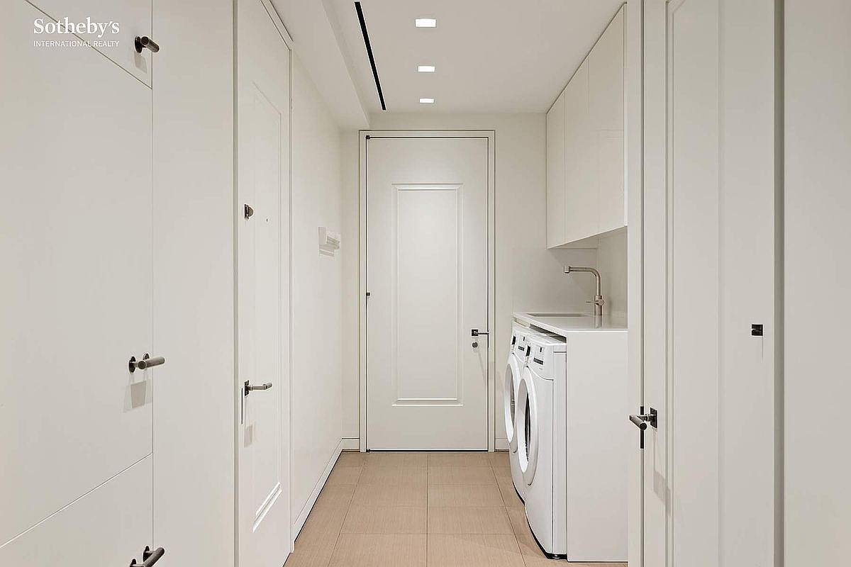 This clean and minimalist laundry room features white cabinetry and a side-by-side washer and dryer set tucked neatly under a counter. The space is finished with light-toned tile flooring and a simple, modern door at the end of the narrow hallway. The bright, neutral color palette creates an organized and functional atmosphere.