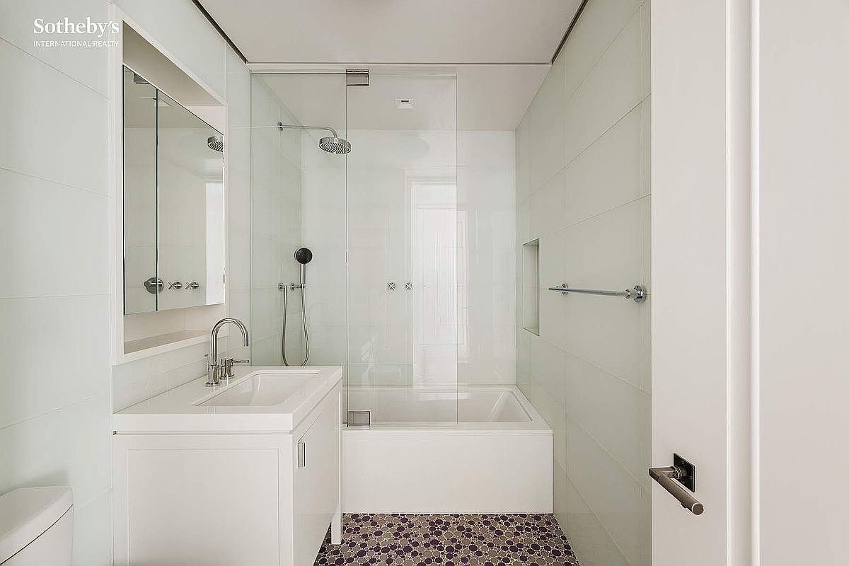 This modern, compact bathroom features a clean white aesthetic with a vanity sink, a glass-enclosed shower-tub combination, and a wall-mounted towel rack. The space is accented by a distinctive dark, circular-patterned tile floor that contrasts with the bright white wall tiles. The perspective is a straight-on view from the doorway, highlighting the efficient layout and contemporary fixtures.