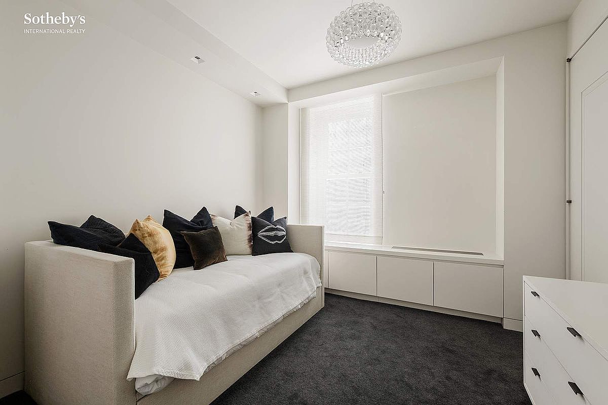 This minimalist guest bedroom features a neutral color palette with a daybed adorned with decorative pillows. A built-in storage unit sits beneath a large window, providing both functionality and a clean, streamlined aesthetic. The room is finished with dark carpeting and a modern, textured light fixture, creating a serene and contemporary atmosphere.