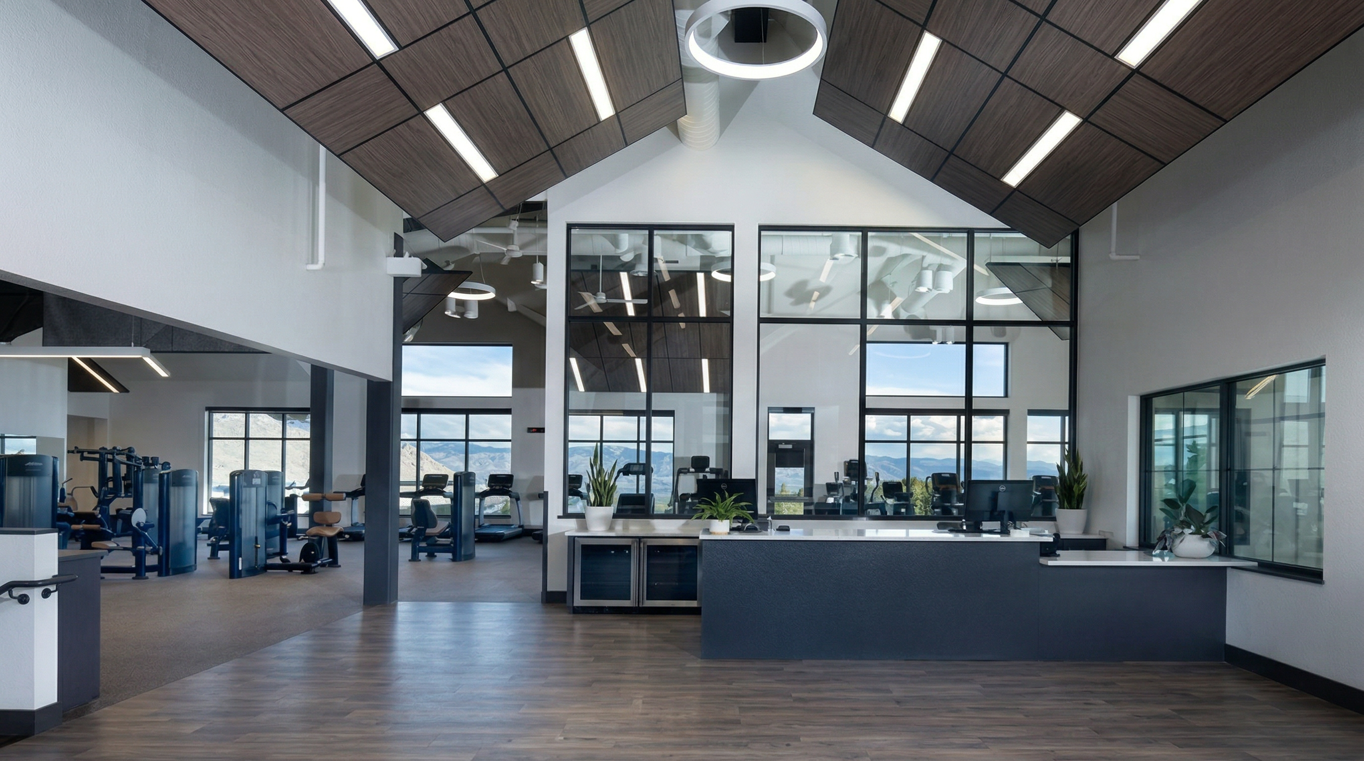 This image showcases a spacious, modern fitness center or clubhouse lobby with high, vaulted ceilings and large windows that flood the space with natural light. The area features a sleek, dark-toned reception desk, wood-look flooring, and a view into an adjacent workout area equipped with various exercise machines. The overall aesthetic is clean, professional, and inviting, highlighting a well-maintained community facility.