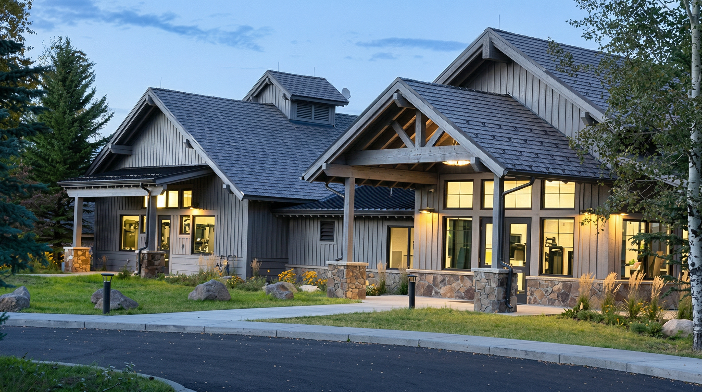 This image captures the exterior front view of a modern, lodge-style community clubhouse or amenity building at dusk. The structure features a combination of vertical wood siding, stone masonry accents, and a multi-gabled roofline, creating a warm and inviting atmosphere. A paved driveway leads up to the entrance, which is framed by a covered portico and illuminated by soft exterior lighting.