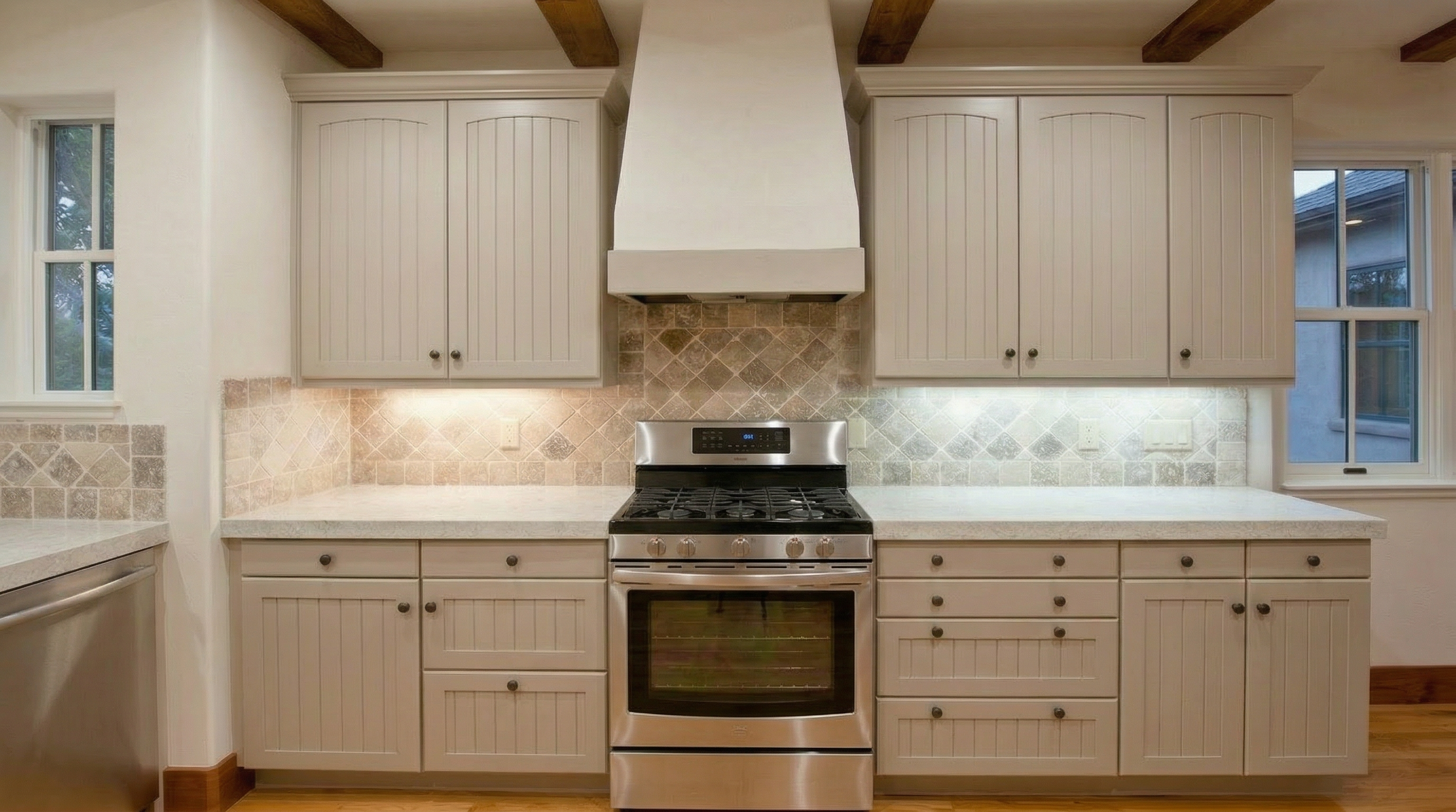 This kitchen features a neutral, classic design with light grey, beadboard-style cabinetry and a prominent custom range hood above a stainless steel double oven. The space is anchored by a diamond-patterned stone tile backsplash and light-colored countertops, creating a warm and cohesive aesthetic. Large windows on either side of the cooking area provide ample natural light, while wooden ceiling beams add a touch of rustic charm to the room.