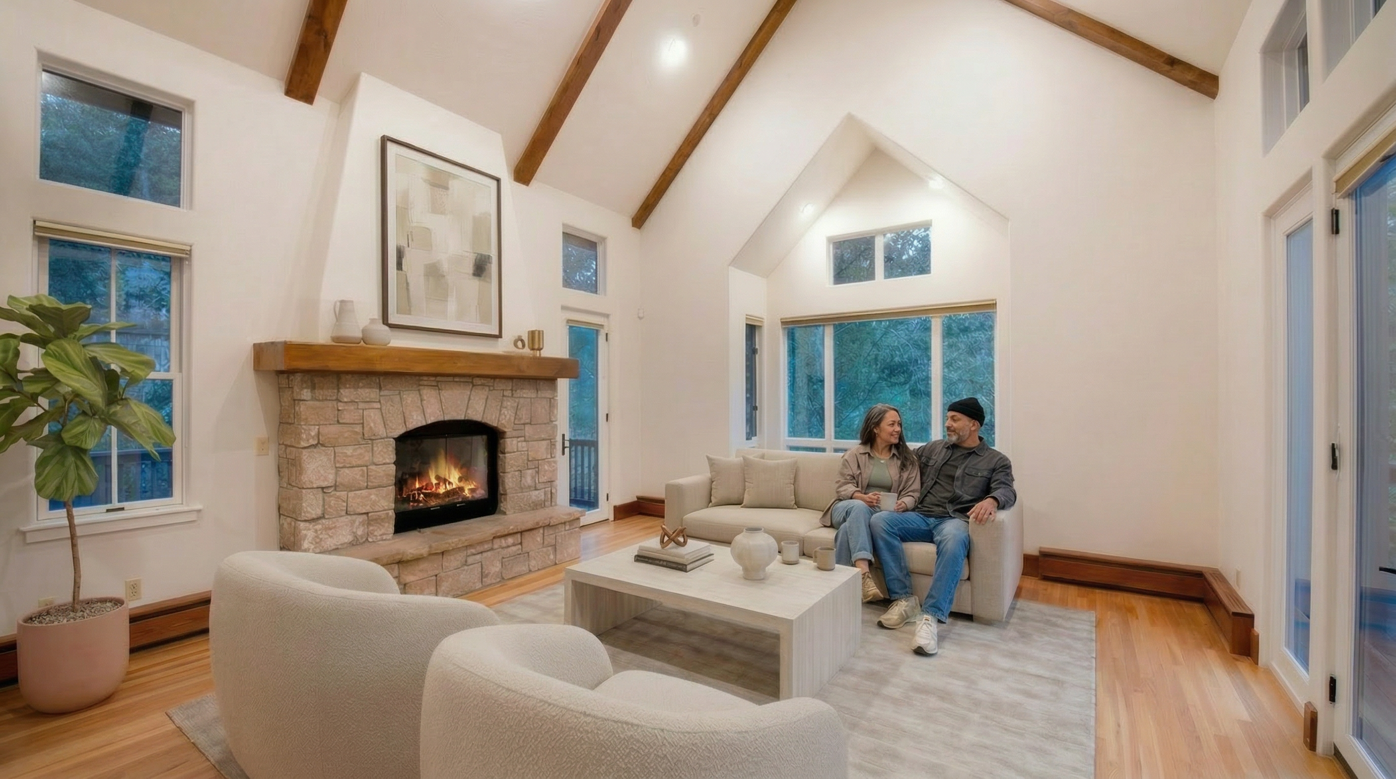 This bright and airy living room features a striking vaulted ceiling with exposed wooden beams, creating a spacious and rustic-modern atmosphere. The focal point is a beautiful stone fireplace with a natural wood mantel, complemented by light oak flooring and neutrally toned furniture. Large windows surrounding the space provide abundant natural light and a connection to the outdoors, highlighting the home's open and inviting layout.