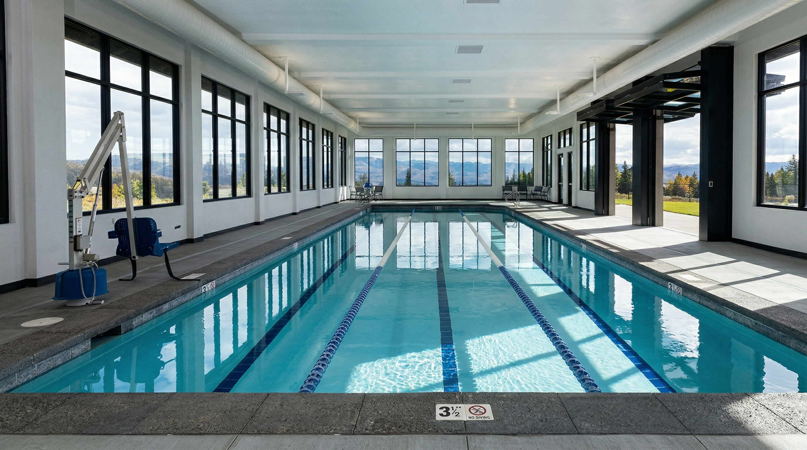 This indoor lap pool features a sleek, modern design with floor-to-ceiling windows that offer expansive views of the surrounding landscape. The space is bright and airy, characterized by clean lines, a neutral color palette, and a professional-grade pool layout with lane markers. It provides a serene and upscale environment, perfect for fitness and relaxation within a residential community.