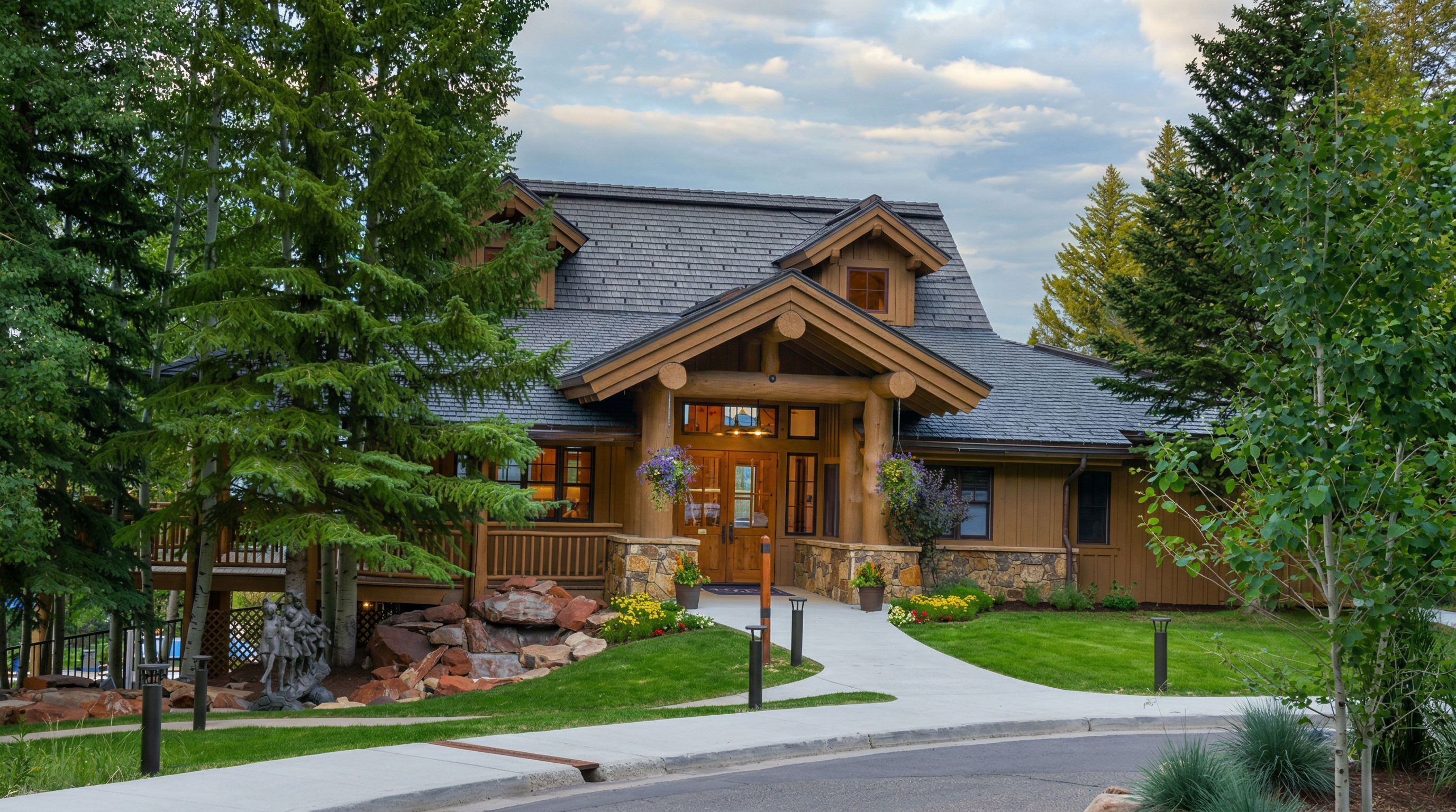 This inviting mountain-style lodge features a prominent timber-framed entryway with natural stone accents and a dark shingled roof. The exterior is surrounded by lush green lawns, mature pine trees, and colorful landscaping, creating a serene and welcoming atmosphere. A paved walkway leads directly to the front doors, emphasizing the property's curb appeal and rustic elegance.