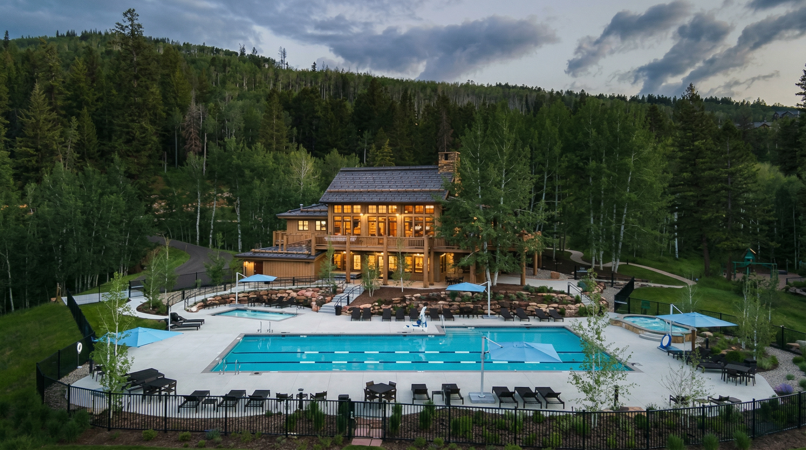 This high-angle aerial shot captures a luxurious mountain lodge-style clubhouse nestled against a dense forest backdrop at dusk. The property features a large, multi-lane swimming pool, a separate hot tub, and expansive stone patio areas furnished with lounge chairs and umbrellas. The warm, inviting glow from the building's interior windows contrasts beautifully with the cool, twilight sky, creating an atmosphere of upscale relaxation and community leisure.