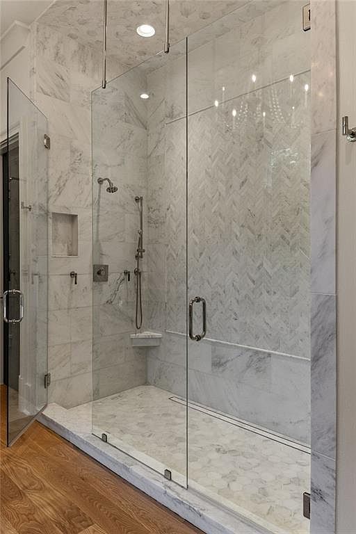 This luxurious walk-in shower features extensive stone tiling in a mix of large-format, herringbone, and hexagonal patterns. The enclosure is defined by clear glass panels and a door with polished metal hardware, complemented by a wall-mounted shower head and handheld sprayer. The perspective is from the adjacent room, showcasing the transition from warm wood flooring to the stone-tiled shower base.