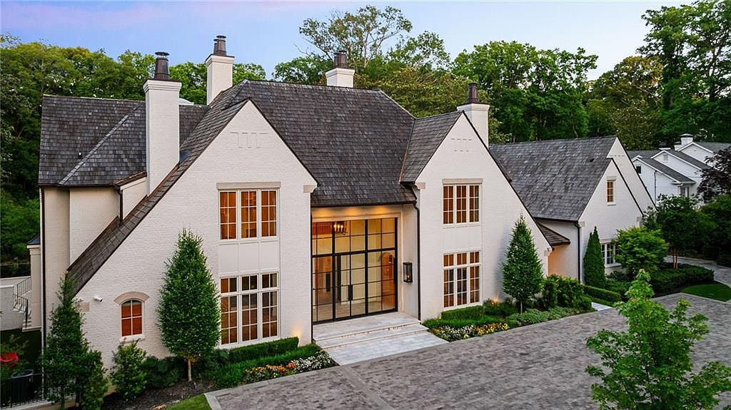 This elegant, multi-gabled estate features a classic white brick facade paired with a dark, textured shingle roof. The central entrance is defined by a striking, large-scale black-framed glass entryway that extends vertically, flanked by symmetrical multi-pane windows. A paved stone driveway leads up to the home, which is surrounded by manicured landscaping and mature trees, creating a sophisticated and welcoming curb appeal.