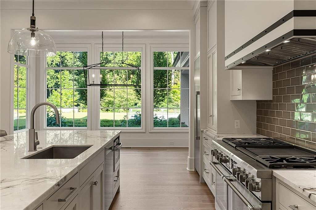 This bright, modern kitchen features a large central island with a stone countertop and an integrated sink, complemented by light-toned cabinetry and a professional-grade stainless steel range. A subway-tiled backsplash adds texture, while a series of large windows provides an expansive view of the lush green outdoors. The space is illuminated by a combination of sleek pendant lighting and recessed fixtures, creating an airy and sophisticated atmosphere.