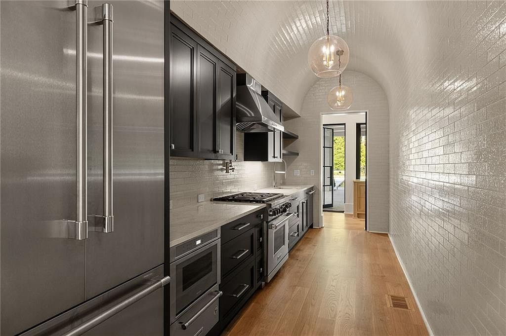 This galley-style kitchen features dark cabinetry contrasted against light stone countertops and a distinctive arched ceiling covered in white subway tile. The space is equipped with stainless steel appliances, including a large refrigerator and a gas range, and is illuminated by two spherical pendant lights. The perspective looks down the narrow corridor toward an open doorway that leads to an outdoor area, highlighting the clean lines and modern aesthetic of the home.