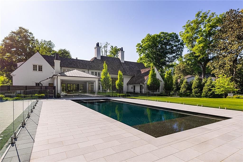 This rear view of a luxury estate showcases a large, rectangular swimming pool with a dark, reflective surface, set within a spacious stone-paved patio. A glass railing borders the pool area, providing an unobstructed view of the manicured lawn and the white, multi-gabled residence in the background. The scene is captured from a low, cinematic angle that emphasizes the clean lines of the architecture and the serene, sunlit outdoor living space.
