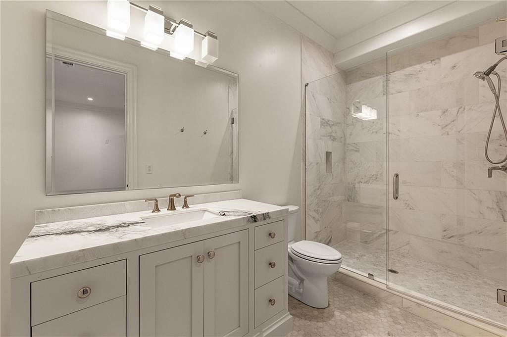 This bright, modern bathroom features a vanity with a stone countertop and a large, rectangular mirror illuminated by a multi-light fixture. To the right, a glass-enclosed shower is lined with light-colored stone tile, while a white toilet sits neatly between the vanity and the shower. The space is finished with neutral-toned cabinetry and patterned floor tile, creating a clean and cohesive aesthetic.