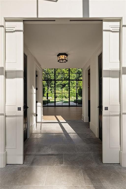 This elegant entryway features a symmetrical design with white paneled double doors opening into a bright, stone-tiled hallway. The space leads the eye toward a large, grid-patterned window wall that frames a lush green outdoor view, creating a seamless transition between indoor and outdoor living. A classic lantern-style light fixture hangs from the ceiling, casting soft shadows across the neutral stone flooring.