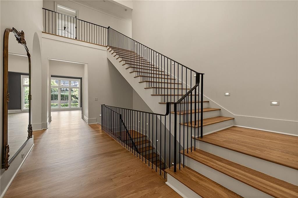 This bright and airy foyer features a grand staircase with light wood treads and a minimalist black metal railing. A large, ornate gold-framed mirror leans against the wall, reflecting the open space and the hallway leading to a windowed room in the background. The neutral walls and light wood flooring create a clean, sophisticated aesthetic, while recessed lighting along the staircase adds a modern touch.
