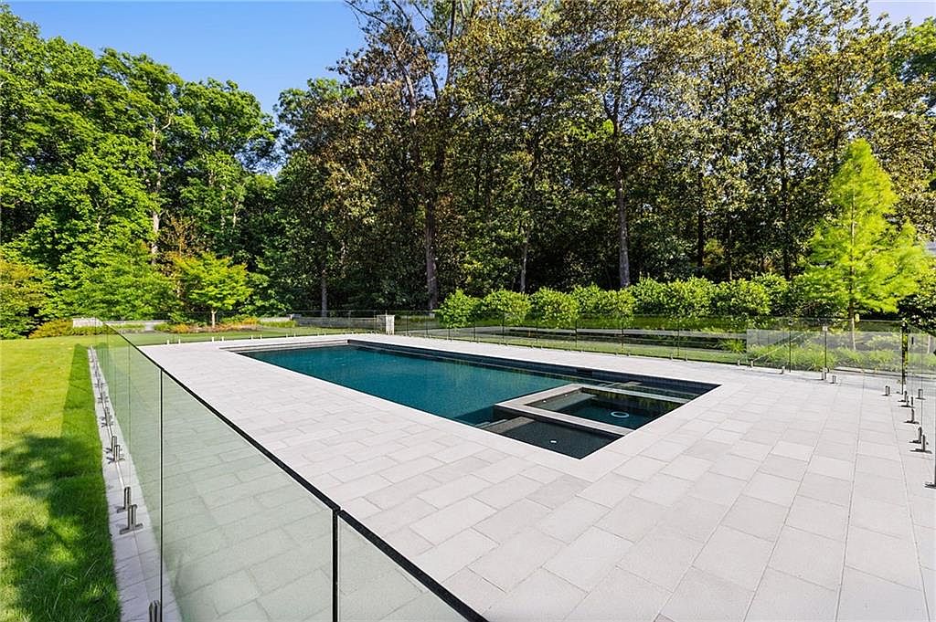 This image captures a sleek, modern rectangular swimming pool integrated with a smaller spa section, set within a spacious light-toned stone patio. A minimalist glass railing surrounds the pool area, providing an unobstructed view of the lush, mature forest backdrop. The scene is bathed in bright, clear daylight, emphasizing the clean lines and tranquil atmosphere of this private outdoor retreat.