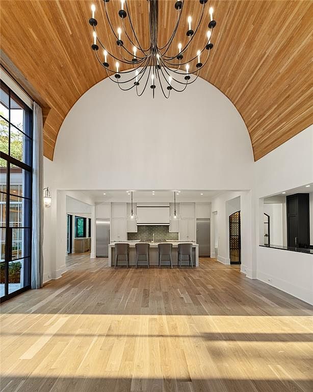 This expansive open-concept kitchen features a dramatic barrel-vaulted ceiling lined with light wood planks and a large, intricate black chandelier. The space centers around a substantial kitchen island with four bar stools, set against a backdrop of white cabinetry and a tiled backsplash. Large, dark-framed windows on the left provide abundant natural light, creating a bright, airy, and sophisticated atmosphere.