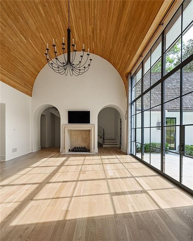 This expansive living room features a dramatic barrel-vaulted ceiling finished in light wood planks and a striking black metal chandelier. A central fireplace with a patterned tile surround serves as a focal point, flanked by two arched doorways leading to other parts of the home. A massive wall of floor-to-ceiling steel-framed windows floods the space with natural light, casting long shadows across the light-toned wood flooring.