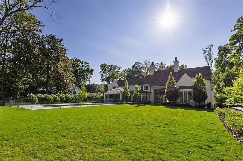 This image captures the expansive rear exterior of a large, traditional-style home set against a bright, sunny sky. The property features a sprawling, well-manicured lawn that leads up to a glass-fenced swimming pool area and a multi-gabled structure with light-colored siding. Tall, mature trees frame the property, creating a private and serene atmosphere perfect for outdoor living.