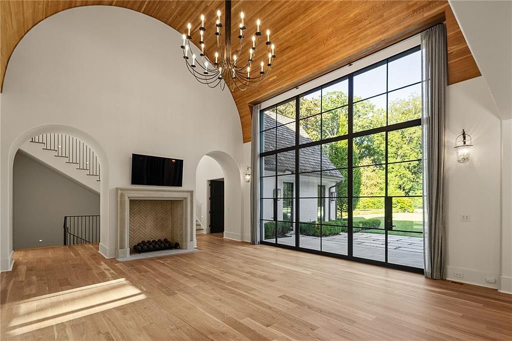 This expansive living room features a dramatic vaulted ceiling with warm wood paneling and a large, multi-pane window wall that provides a clear view of the exterior grounds. A stone fireplace with a herringbone-patterned firebox serves as a central focal point, complemented by a grand chandelier suspended from the arched ceiling. The space is finished with light wood flooring and arched doorways that lead to adjacent areas, creating an airy and sophisticated atmosphere.