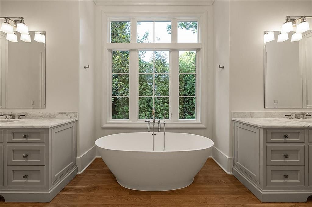 This primary bathroom features a symmetrical layout with a central freestanding soaking tub positioned beneath a large, multi-pane window that overlooks greenery. Flanking the tub are dual vanities with light-toned cabinetry, stone countertops, and wall-mounted mirrors with matching light fixtures. The room is finished with warm-toned wood flooring, creating a clean, balanced, and serene atmosphere.