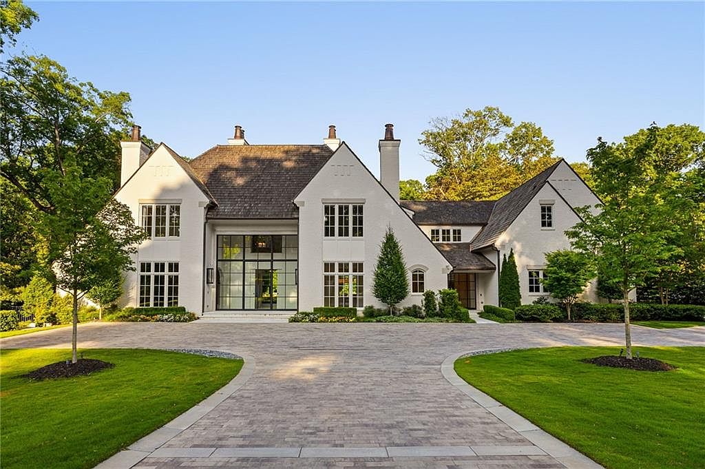 This grand, white-painted brick residence features a symmetrical architectural design with steep gabled rooflines and a prominent central glass entryway. A wide, paved circular driveway leads up to the home, framed by manicured green lawns and mature trees. The exterior aesthetic is sophisticated and timeless, showcasing a blend of traditional structure with modern, large-scale glazing.