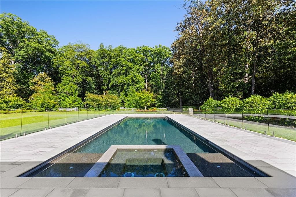 This image captures a sleek, rectangular swimming pool with an integrated square spa in the foreground, set against a backdrop of lush, mature trees. The pool area is surrounded by light-toned stone pavers and enclosed by a modern glass fence, creating a seamless transition to the expansive green lawn beyond. The perspective is low and centered, emphasizing the clean lines and tranquil atmosphere of this private outdoor oasis.
