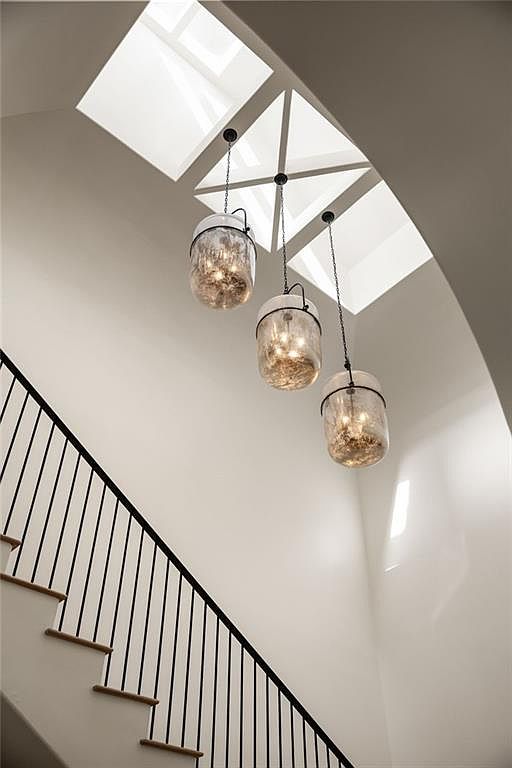 This image captures a bright, modern staircase area featuring a clean, minimalist aesthetic with white walls and a black metal railing. Three decorative glass pendant lights with textured interiors hang at varying heights, drawing the eye upward toward a geometric skylight arrangement that floods the space with natural light. The perspective is looking up from the staircase, emphasizing the verticality and architectural interest of the light-filled stairwell.