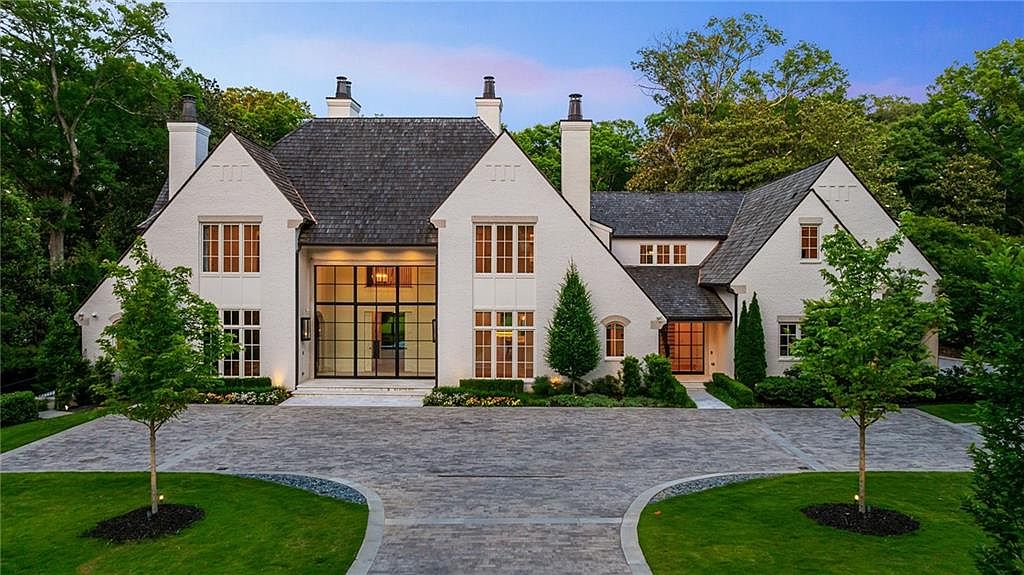 This grand, white-painted brick estate features a classic architectural design with a steep, dark-shingled roof and multiple chimneys. The central entrance is defined by a striking, multi-paned glass wall that spans two stories, flanked by symmetrical window arrangements. A wide, paved motor court leads up to the home, framed by manicured lawns and mature trees, creating an elegant and welcoming curb appeal.