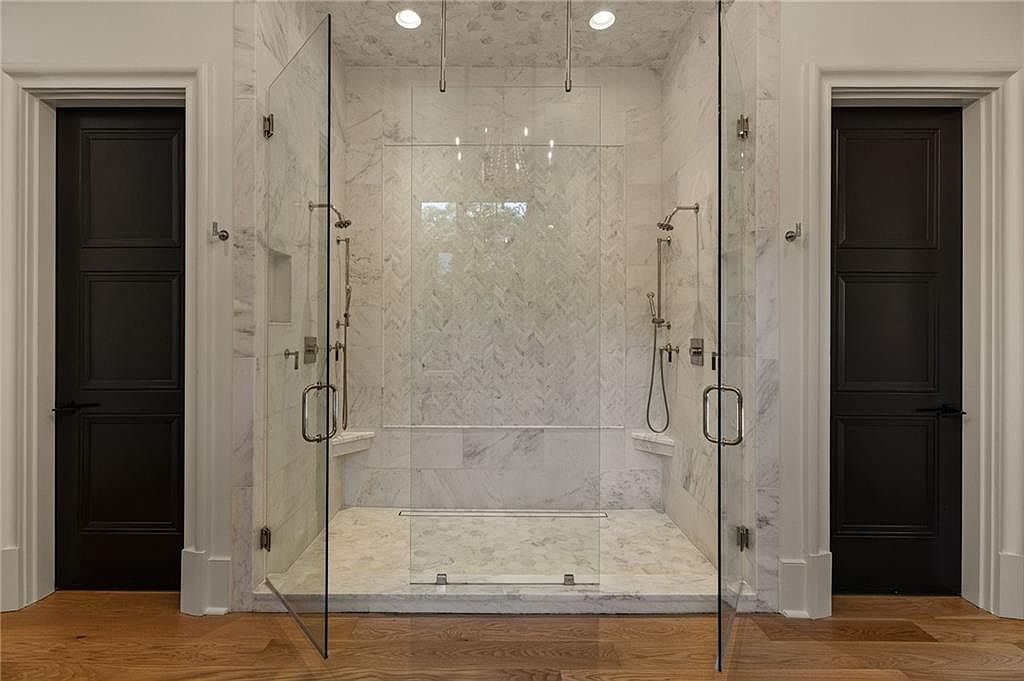 This primary bathroom features a spacious walk-in shower enclosed by glass panels, showcasing intricate stone tile work in a herringbone pattern on the back wall. The shower includes dual showerheads, built-in corner benches, and a linear drain, flanked by two dark-toned doors that provide a striking contrast to the light stone surfaces. The perspective is centered and symmetrical, highlighting the high-end finishes and clean, modern aesthetic of the space.