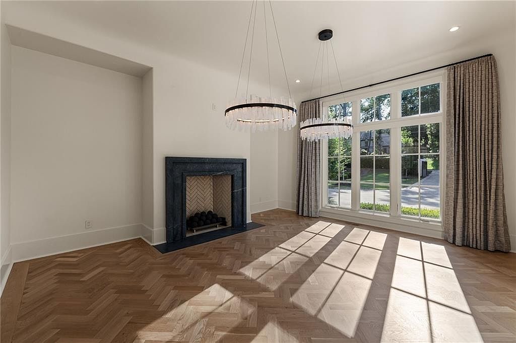 This elegant living room features light wood flooring laid in a classic herringbone pattern and a dark stone fireplace surround with a herringbone-patterned firebox interior. Two modern, circular crystal chandeliers hang from the ceiling, casting a sophisticated glow over the space. Large windows with floor-length curtains allow natural light to stream in, creating a bright and airy atmosphere with a view of the outdoors.
