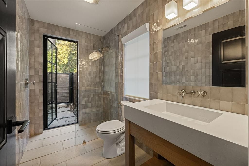 This modern bathroom features a floating vanity with a light wood base and a thick, white countertop paired with wall-mounted faucets. The space is finished with textured, neutral-toned square tiles on the walls and a glass-enclosed shower area that opens directly to an outdoor patio through a black-framed door. The perspective captures the clean lines of the vanity and the seamless transition between the indoor shower and the exterior space.
