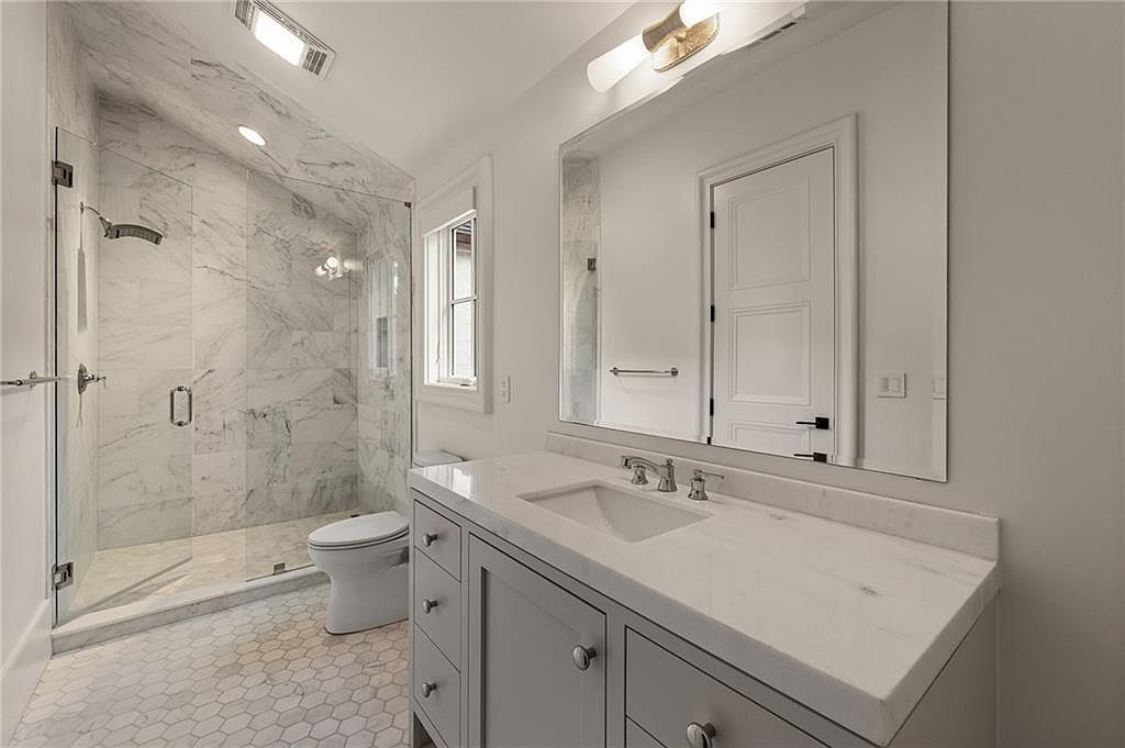 This bright, modern bathroom features a vanity with a light-toned stone countertop and gray cabinetry, paired with a large wall-mounted mirror. To the left, a walk-in shower is enclosed by glass panels and finished with light-colored, veined stone tile. The floor is covered in a classic hexagonal tile pattern, and the space is illuminated by a sleek, multi-bulb vanity light fixture.