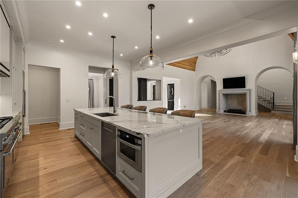 This spacious, open-concept kitchen features a large central island with a stone countertop, integrated appliances, and bar seating. The room flows seamlessly into a living area characterized by light wood flooring, arched architectural details, and a stone-clad fireplace. The perspective captures the transition between the functional culinary space and the expansive living area, highlighting the bright, airy atmosphere created by recessed lighting and neutral tones.