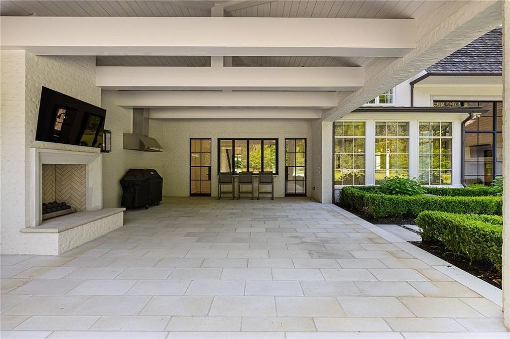 This expansive covered patio features a light-toned stone floor and a white-painted brick structure with exposed beams overhead. The space is equipped with an outdoor fireplace, a built-in grill area, and a bar-style seating arrangement looking out toward the manicured garden. The design creates a seamless transition between indoor and outdoor living, offering a sophisticated and airy atmosphere for entertaining.