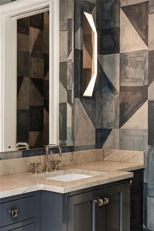 This sophisticated bathroom vanity area features a dark, moody cabinet base topped with a light-toned stone countertop and an integrated sink. The wall is adorned with a striking geometric patterned wallpaper in shades of blue, gray, and cream, complemented by a sleek, modern vertical wall sconce. A mirror reflects the patterned wall, adding depth to the space and creating a polished, high-end aesthetic.