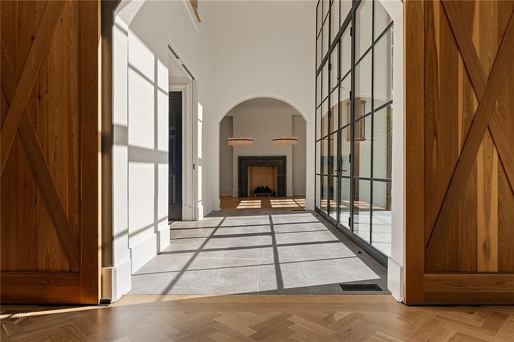 This elegant hallway features a striking transition from light wood herringbone flooring to large-format stone tiles. The space is defined by a high, arched opening leading to a distant room with a fireplace, while a wall of floor-to-ceiling glass panels on the right allows natural light to cast dramatic grid-patterned shadows across the floor. Large wooden barn-style doors frame the foreground, creating a sophisticated and architectural entryway into the home's interior.