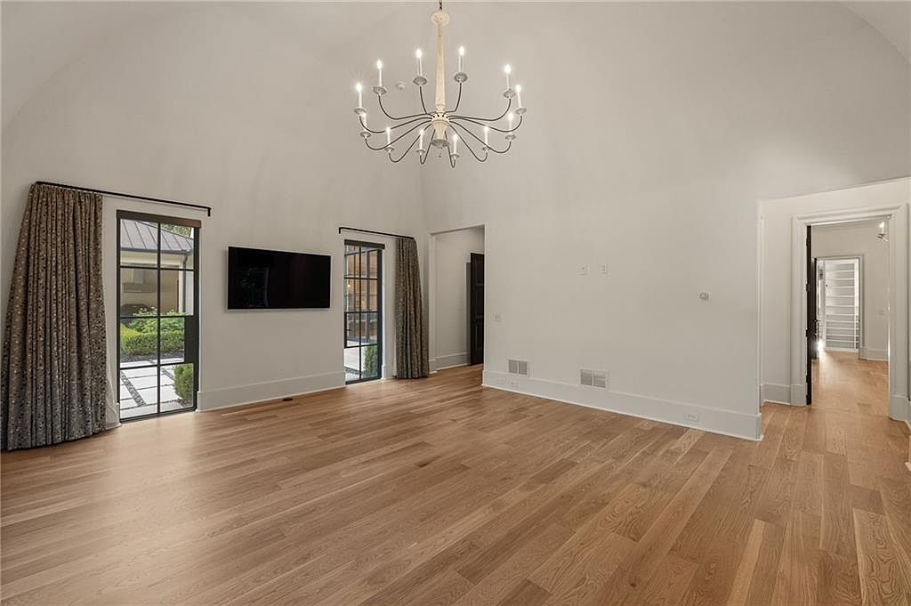 This spacious living room features light wood flooring and a high, vaulted ceiling accented by a large, multi-armed chandelier. Two sets of tall windows with dark frames and patterned curtains provide views of the exterior, while a wall-mounted television serves as a focal point. The room has a clean, neutral aesthetic with a doorway leading to an adjacent hallway, creating an open and airy atmosphere.