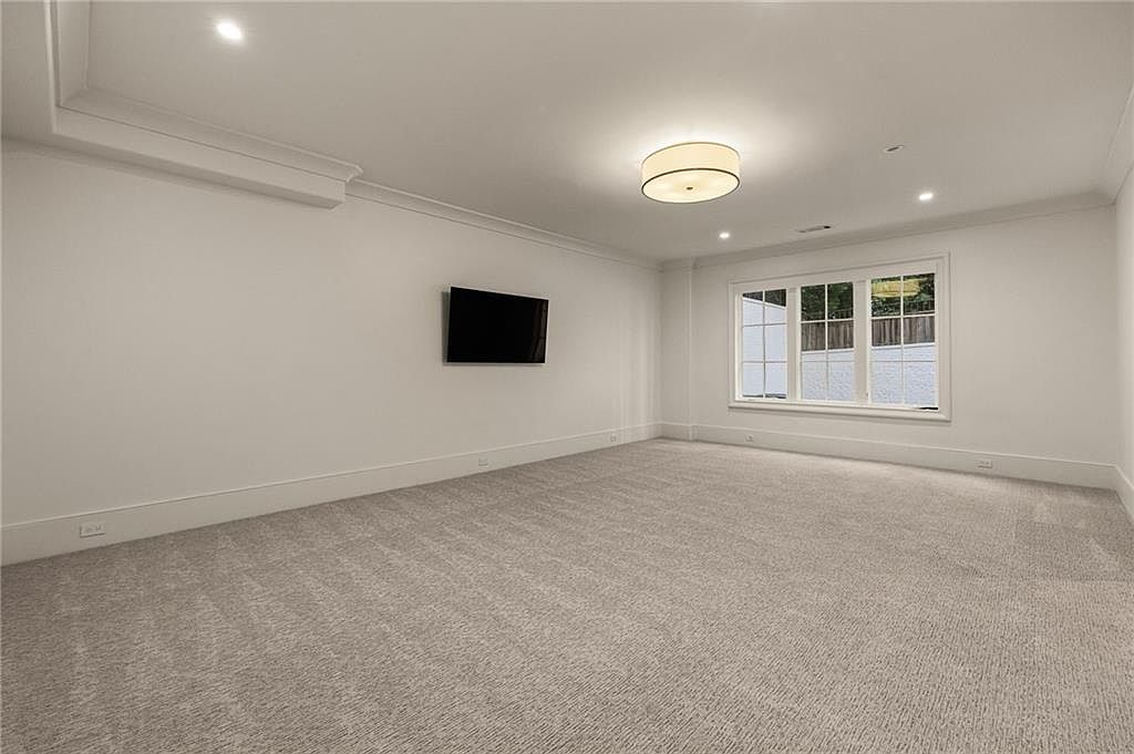 This spacious, minimalist living area features neutral-toned carpeting and crisp white walls, creating a bright and airy atmosphere. A wall-mounted television serves as a focal point, while a large, multi-pane window allows natural light to fill the room. The space is illuminated by a central drum-style ceiling fixture and recessed lighting, offering a clean and versatile canvas for interior design.