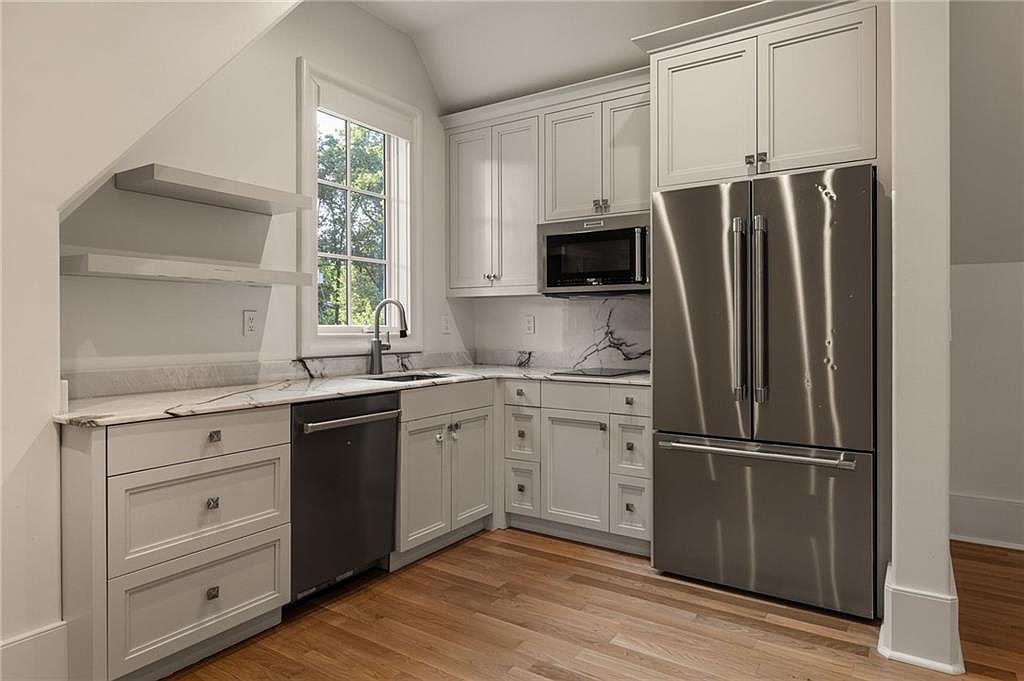 This compact, modern kitchen features light-colored cabinetry, stainless steel appliances, and stone countertops with a subtle veined pattern. The space is efficiently designed with an L-shaped layout, including a built-in microwave, a dishwasher, and a large refrigerator, all set against a backdrop of light wood flooring. A window above the sink provides natural light, while open shelving adds a decorative touch to the angled wall.