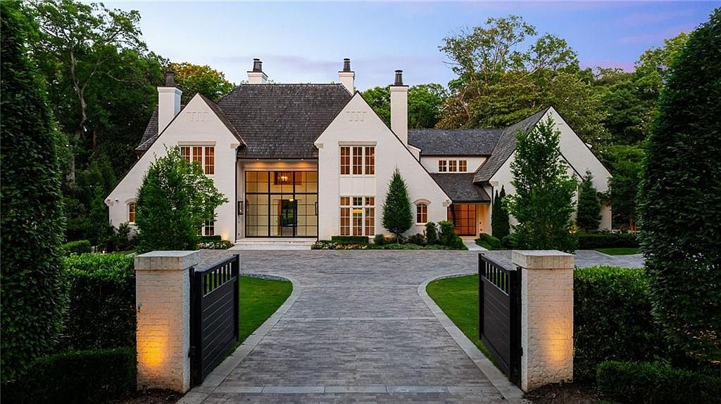 This grand, white-painted brick estate features a symmetrical architectural design with steep, dark-shingled rooflines and multiple chimneys. A paved driveway leads through a gated entrance toward a striking, large-scale glass-paned entryway that serves as the focal point of the facade. The property is framed by manicured hedges and mature trees, creating a sophisticated and private curb appeal.