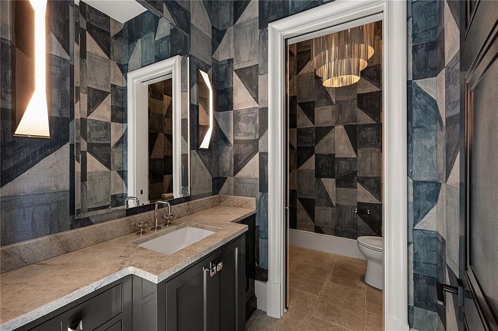This sophisticated powder room features bold, geometric patterned wallpaper in shades of blue and grey, creating a striking visual impact. The vanity is topped with a neutral stone counter and an undermount sink, complemented by modern wall-mounted light fixtures and a large mirror. A doorway leads into a separate water closet area, which is illuminated by a decorative tiered chandelier and features matching wallpaper.