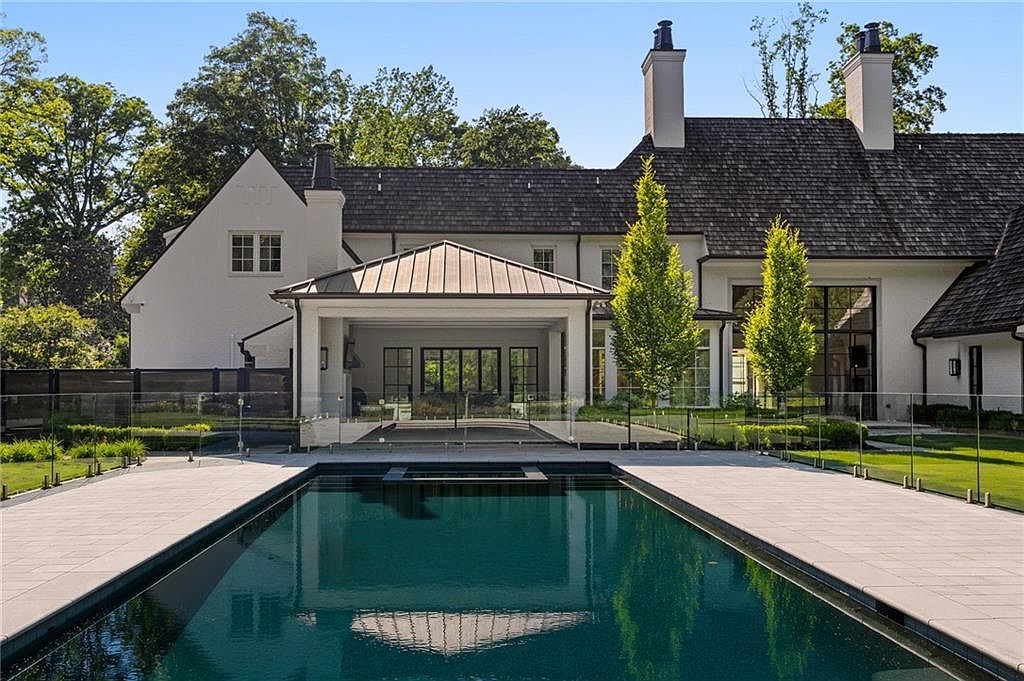 This rear view of a luxury residence showcases a pristine, dark-bottom swimming pool with a built-in spa, framed by light-toned stone decking and a modern glass safety fence. The home features a white stucco facade, a dark shingled roof, and a covered outdoor living area with a metal roof, creating a sophisticated and serene atmosphere. Two slender, vibrant green trees stand symmetrically in the manicured lawn, enhancing the elegant and well-maintained aesthetic of the backyard.