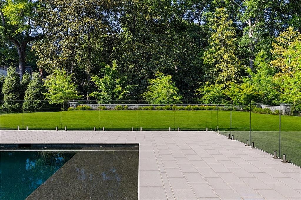 This image captures a serene outdoor living space featuring a dark-tiled swimming pool adjacent to a light-colored stone patio. A modern glass railing separates the paved area from a lush, manicured green lawn that stretches toward a dense backdrop of mature trees. The perspective is from the poolside, emphasizing the clean lines and contemporary design of the backyard landscape.