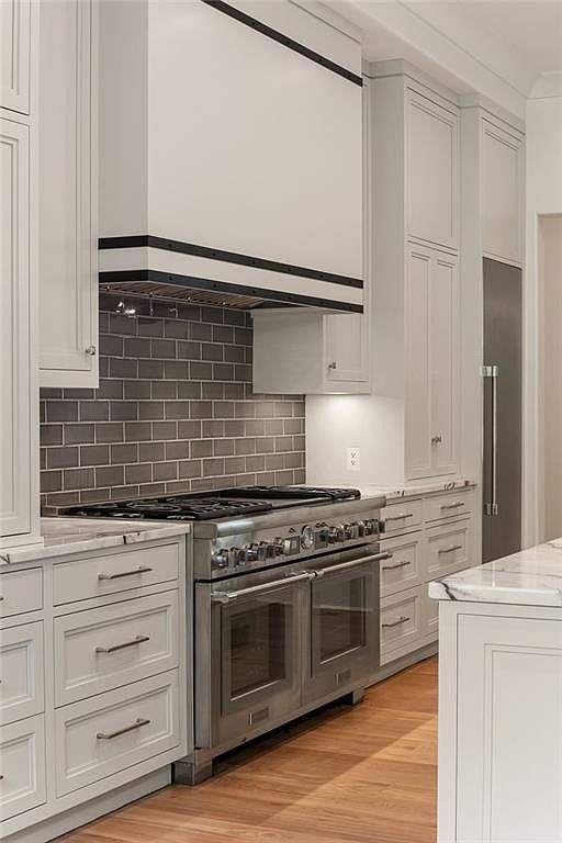 This kitchen features a professional-grade stainless steel range set against a dark subway tile backsplash. A custom-built range hood with dark metal accents serves as a focal point above the stove, flanked by light-colored cabinetry. The space is finished with light wood flooring and stone countertops, creating a bright and sophisticated culinary environment.